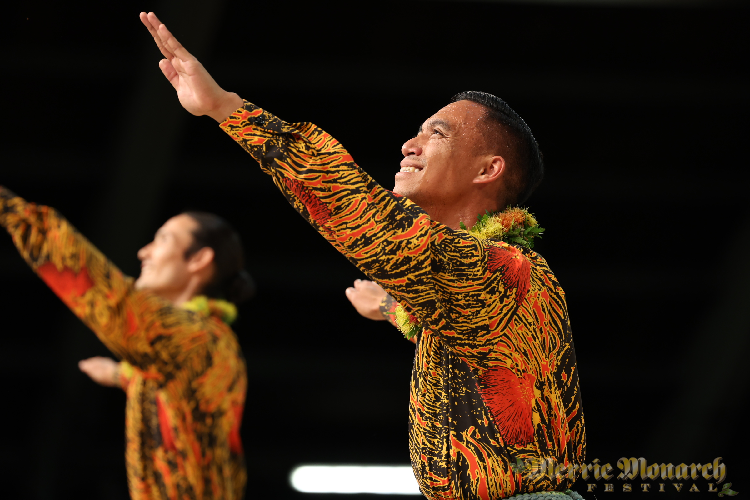 Kane hula dancers wearing black with red and yellow lehua prints and flower lei.